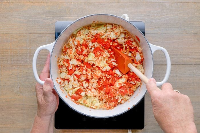 A person stirs chopped cabbage and tomatoes in a large white pot on a stovetop with a wooden spoon.