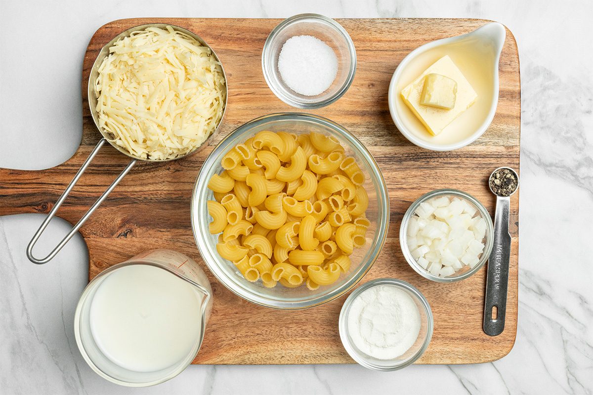 A wooden board holds bowls of dry pasta, shredded cheese, diced onions, butter, salt, pepper, flour, and a pitcher of milk, all arranged neatly for cooking preparation.