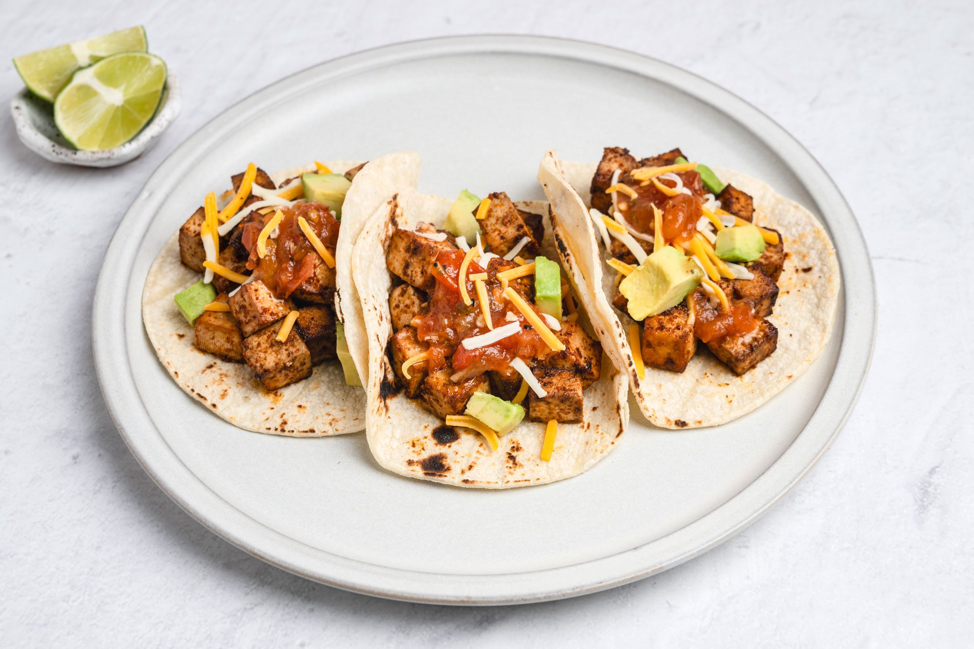 3/4th shot of Three tortillas filled with seasoned tofu, avocado cubes, salsa, and shredded cheese are arranged on a round plate, A small dish with lime wedges is placed in the background