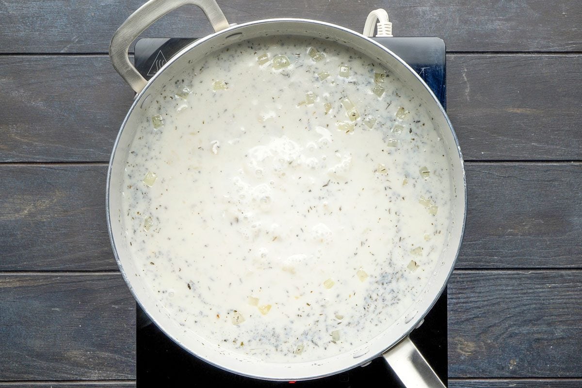 Top Shot of a silver saucepan with creamy white sauce; herbs; and small chunks simmers on a stovetop; viewed from above against a dark wooden background, creating contrast and highlighting the rich texture of the simmering mixturex
