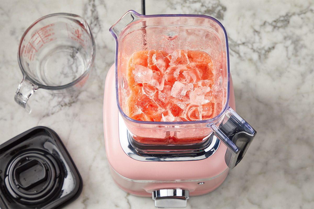 Top-down view of a pink blender filled with ice and pieces of watermelon on a marble countertop. Next to it is an empty measuring cup and the blender lid.