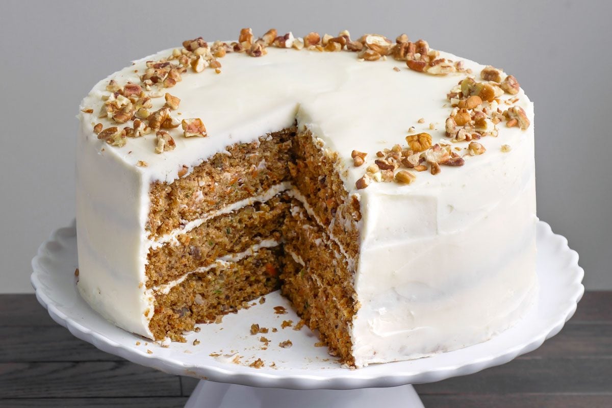 3/4 angle view of Zucchini Carrot Cake on a cake tray; A large slice has been removed; arranged on a dark brown wooden surface;