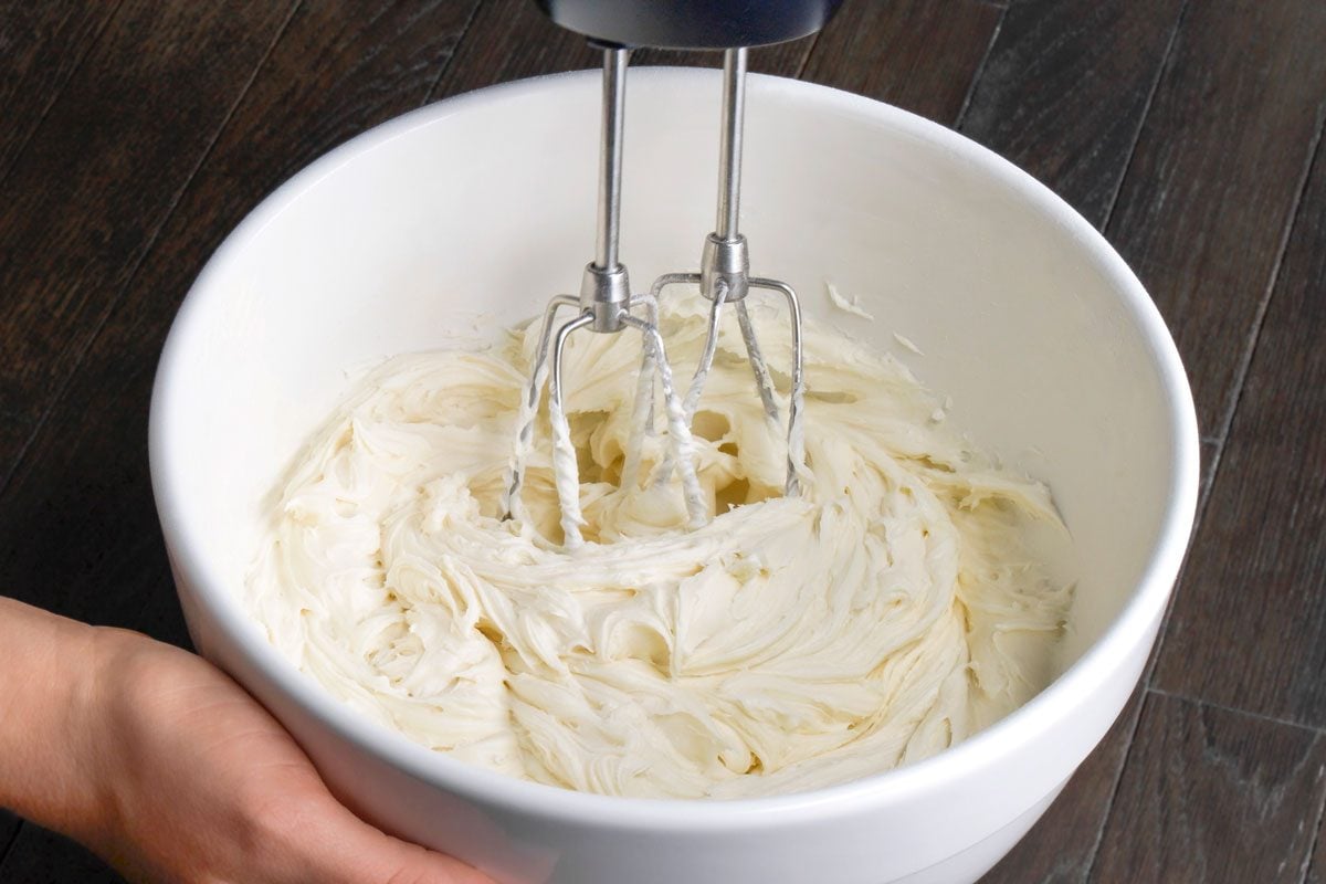 Close shot of a large bowl; For frosting beat cream cheese and butter in a large bowl until smooth; Add sugar and vanilla; Continue beating until sugar is dissolved; hand blender; all set on adark brown wooden surface;