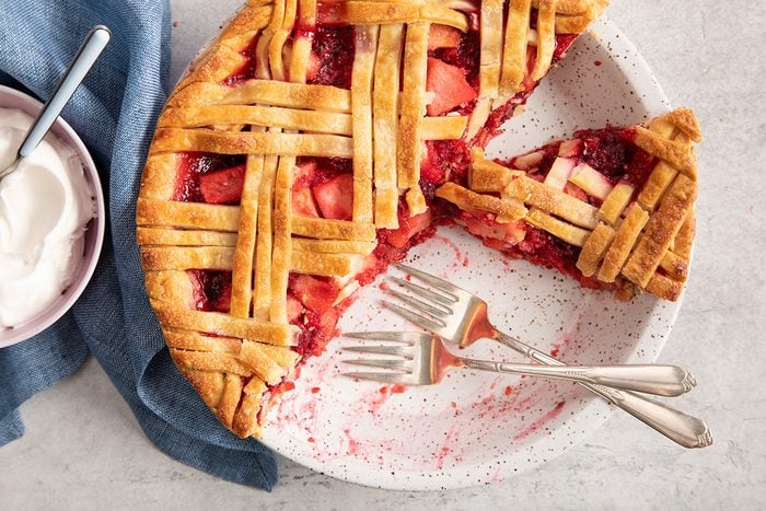 A lattice-topped fruit pie with a slice removed sits on a speckled plate, accompanied by three forks. A bowl of whipped cream and a blue napkin are nearby.
