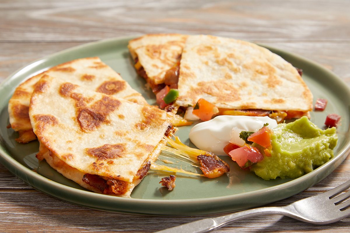 A green plate with cheese quesadillas cut into wedges, served with a side of guacamole, sour cream, and diced tomatoes. A fork is placed on the table next to the plate.