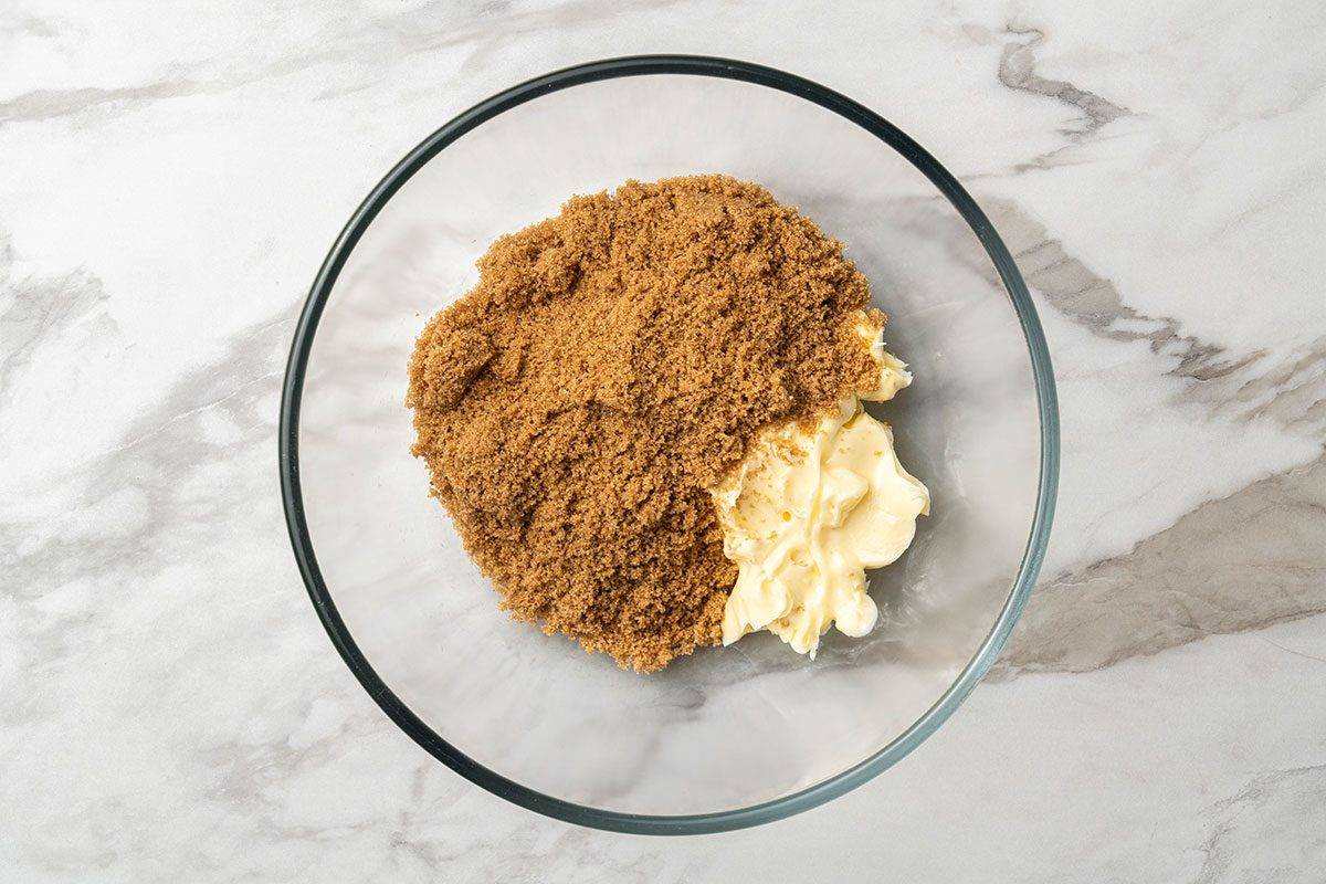 A clear glass bowl on a marble surface contains light brown sugar and a portion of soft, pale yellow butter placed side by side.