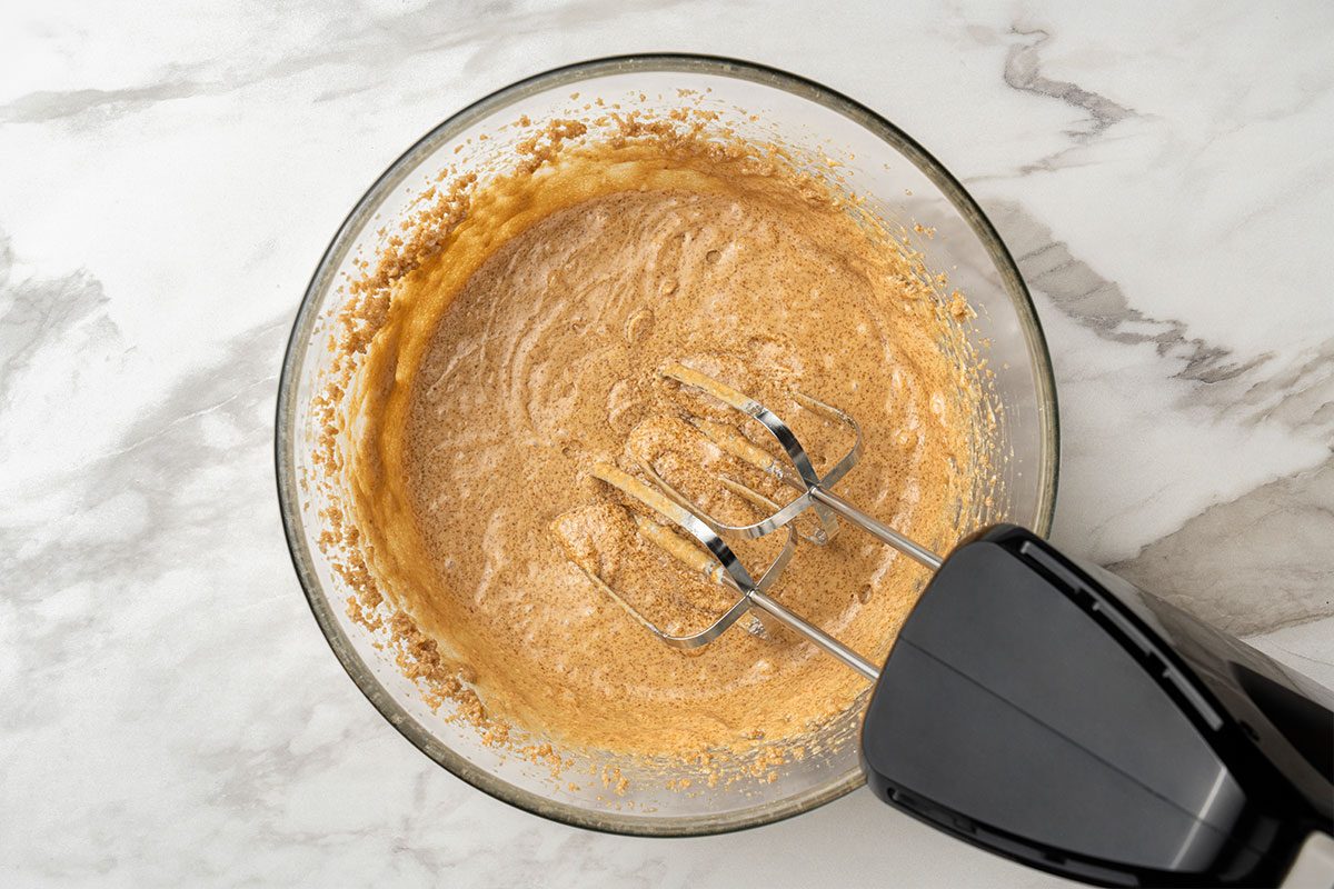 A glass bowl filled with a light brown batter is being mixed with a black electric hand mixer on a white marble countertop.