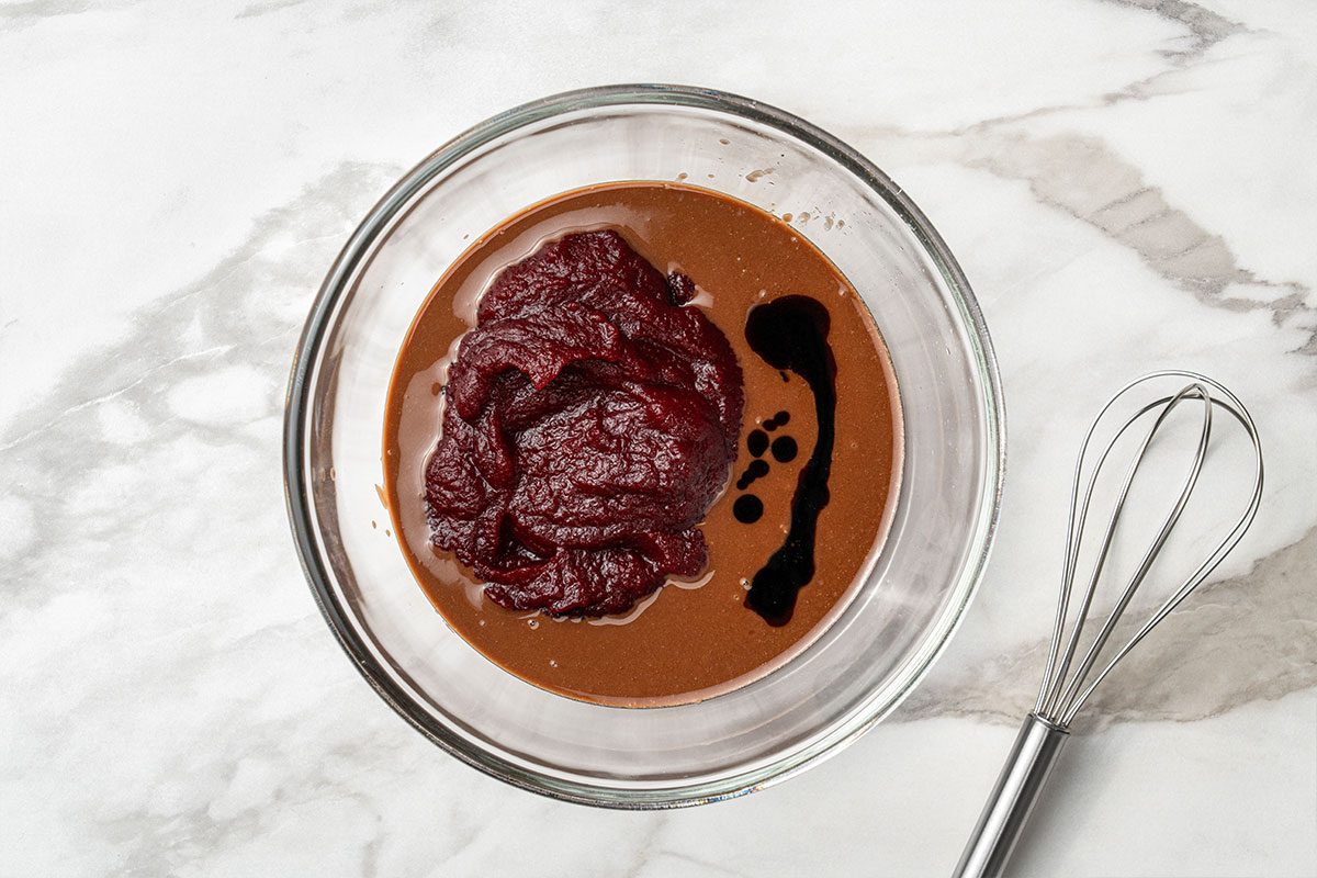 A glass bowl containing tomato paste, a dark liquid, and a brown sauce sits on a white marble surface next to a metal whisk.