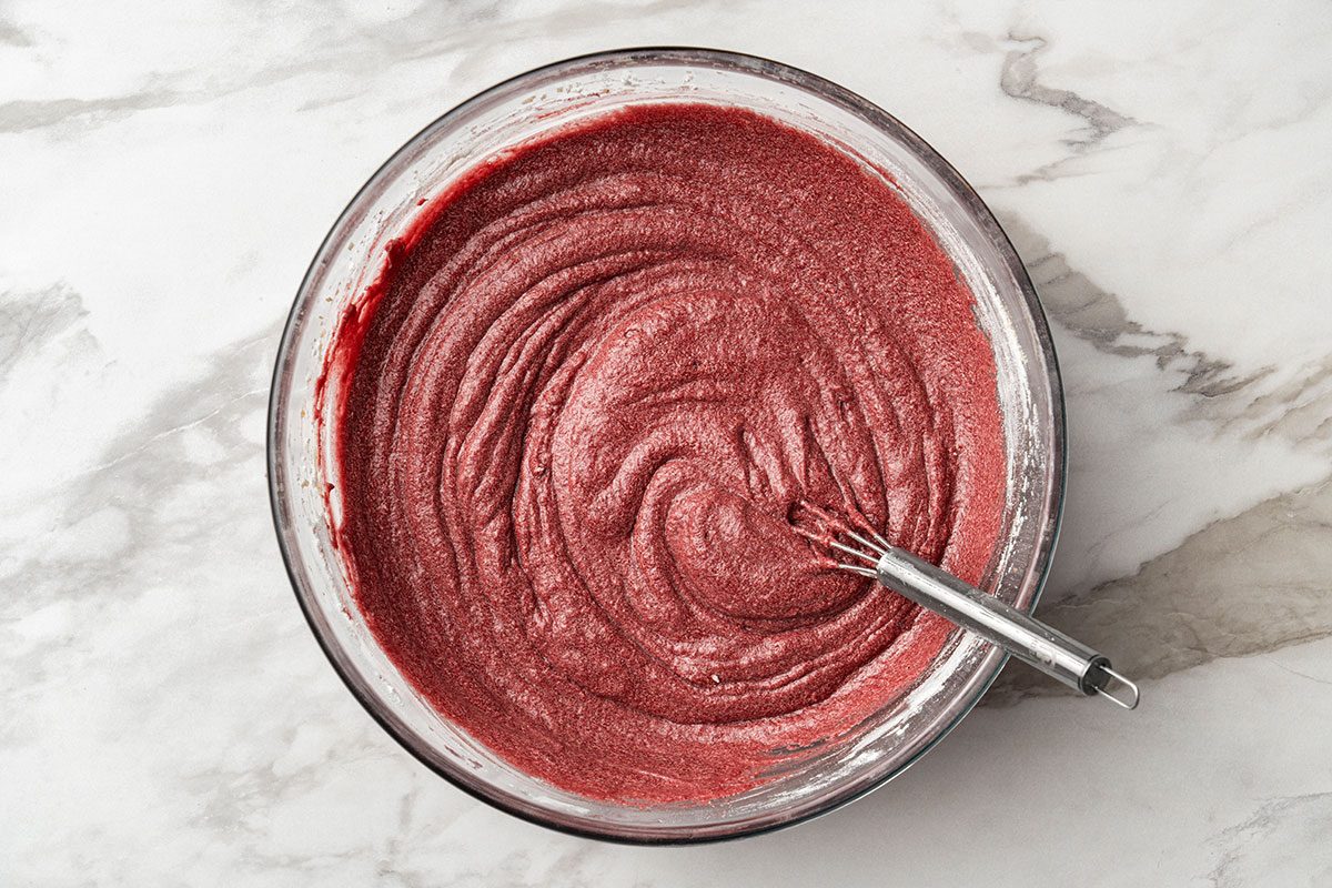 A glass bowl filled with red cake batter being mixed with a metal whisk, placed on a white marble countertop.
