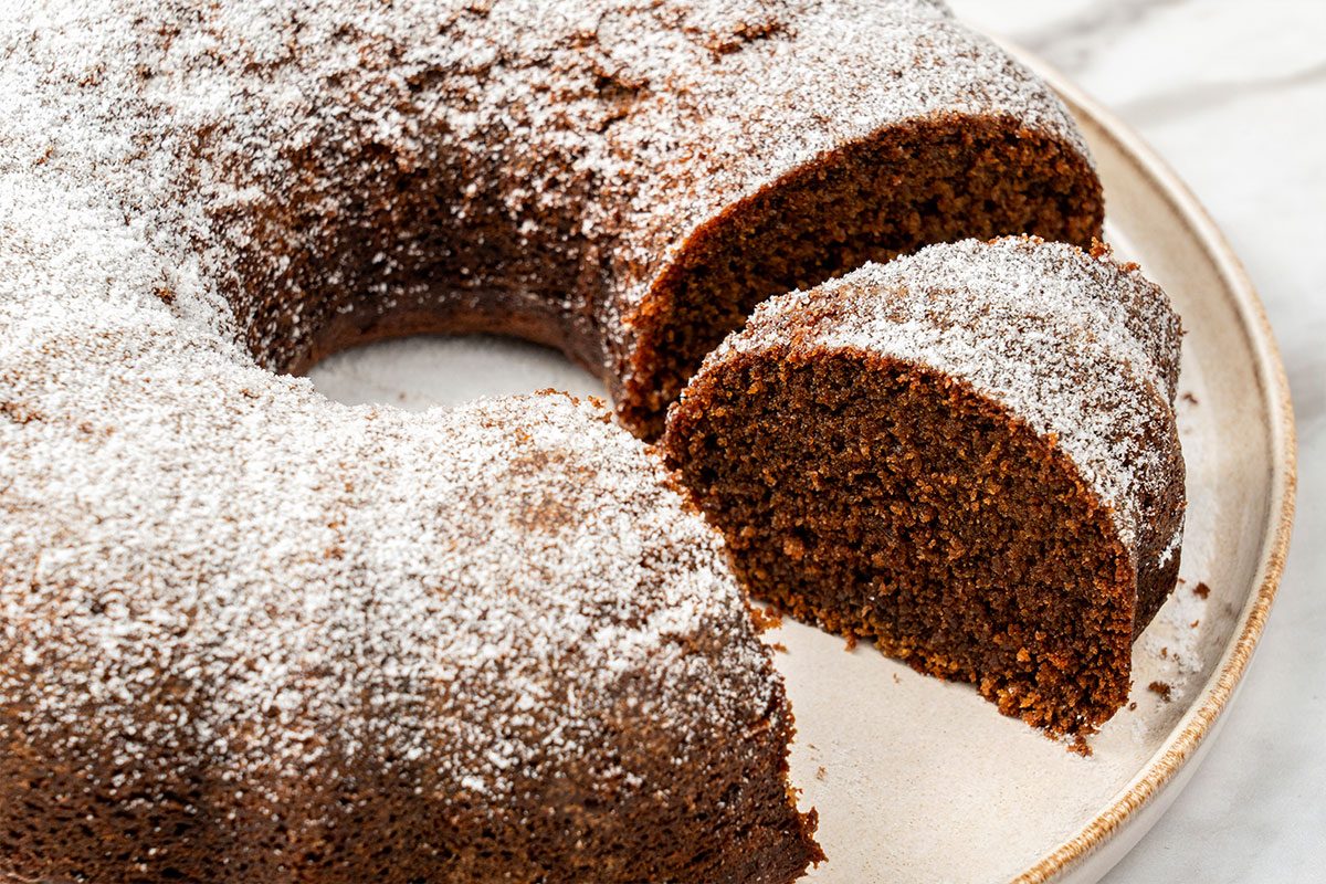 A round chocolate bundt cake lightly dusted with powdered sugar sits on a plate, with a slice partially cut and slightly separated from the cake.