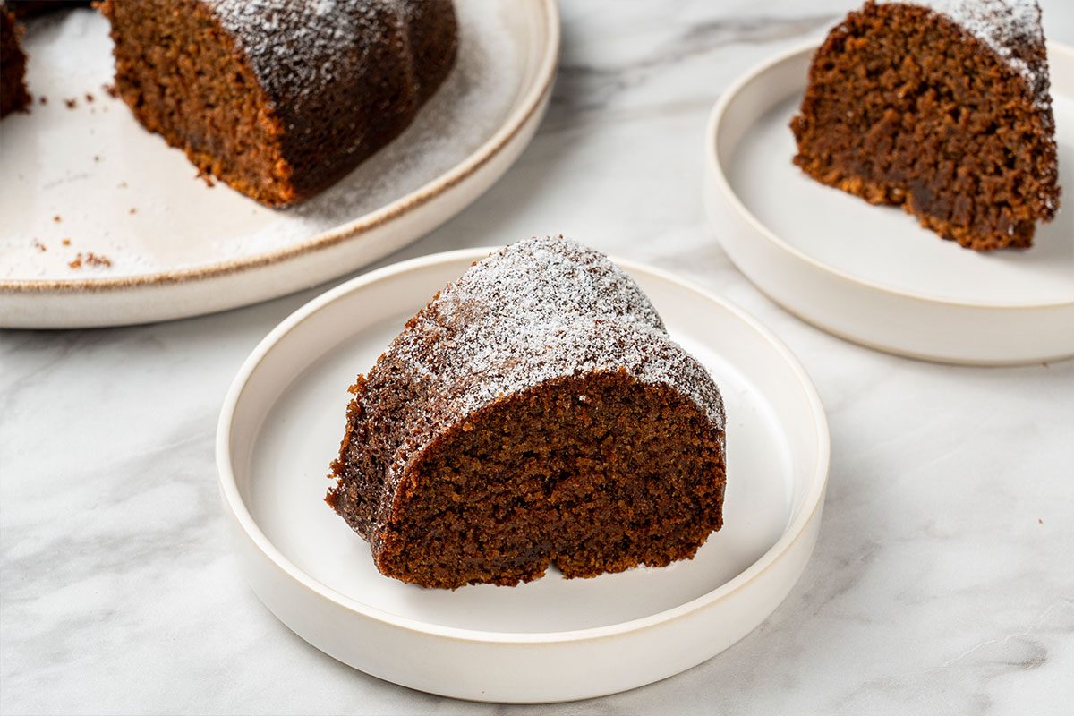 A slice of chocolate bundt cake topped with powdered sugar is served on a white plate, with more cake slices visible on plates in the background.