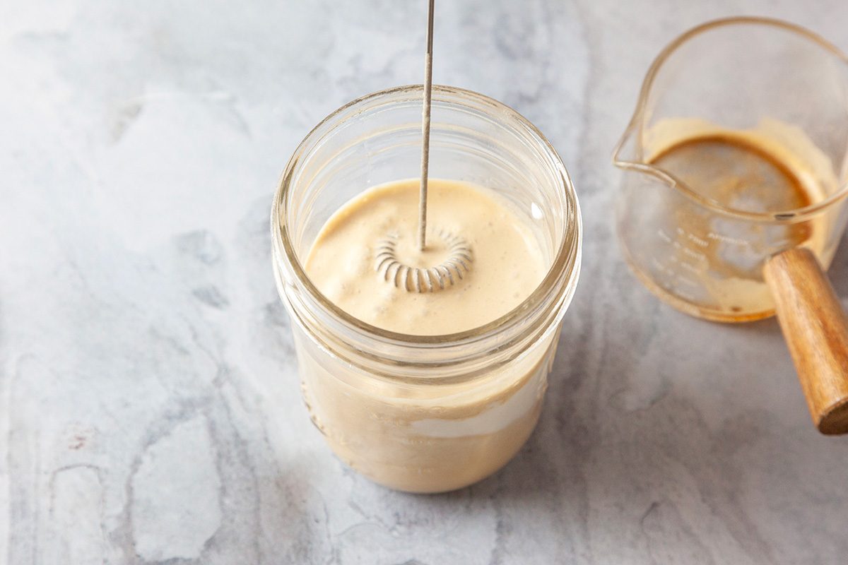 Overhead shot of a handheld milk frother whips creamy coffee in a glass jar; next to it is an empty glass measuring cup with coffee residue and a wooden handle on a light countertop