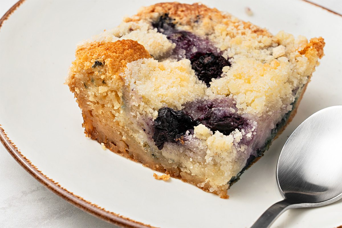A close-up of a slice of blueberry coffee cake on a white plate, with a silver spoon beside it. The cake has a crumbly topping and visible blueberries baked inside.