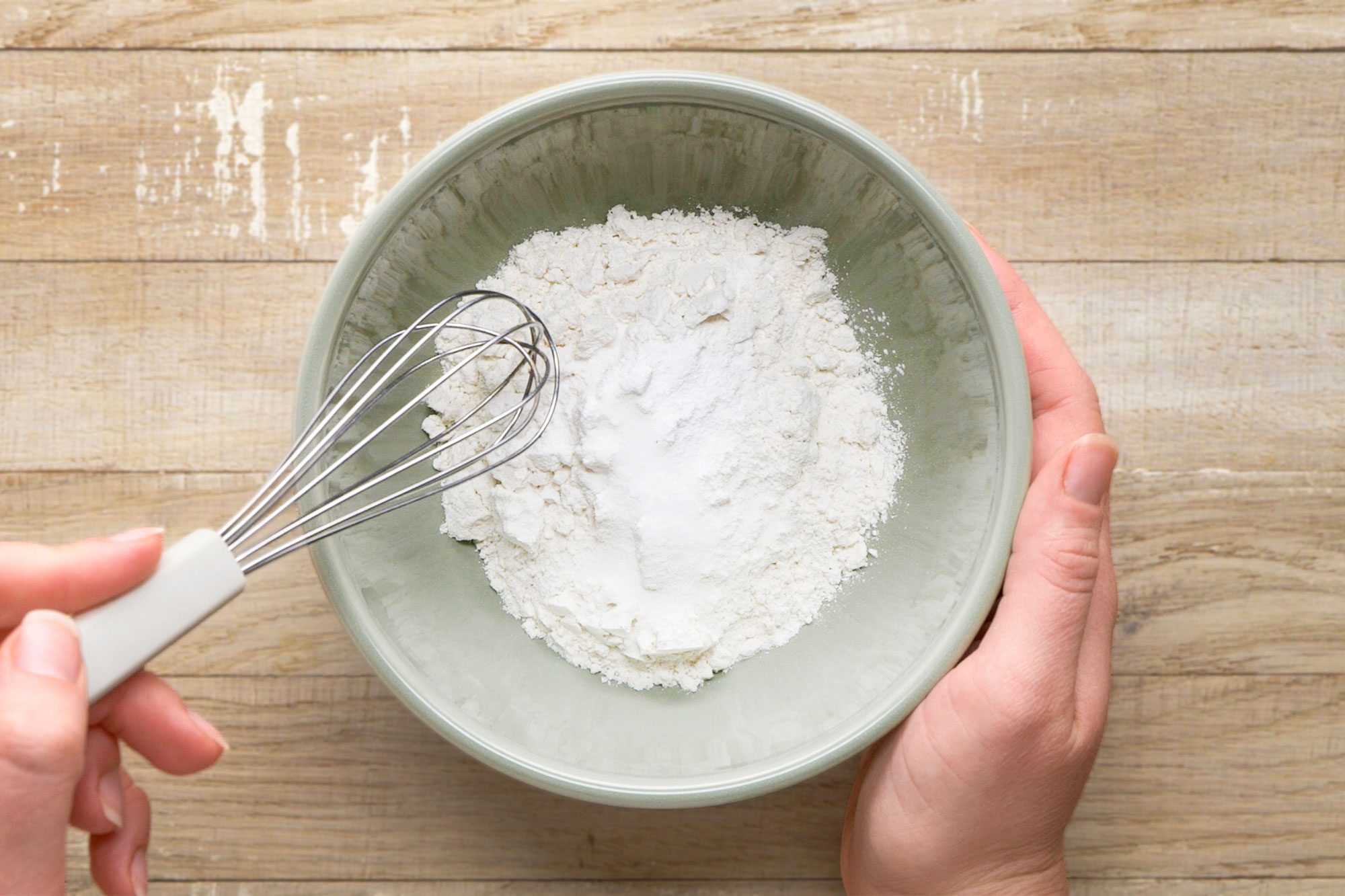 overhead shot of a person holding a green bowl filled with flour and baking powder, mixing the ingredients with a metal whisk on a wooden surface