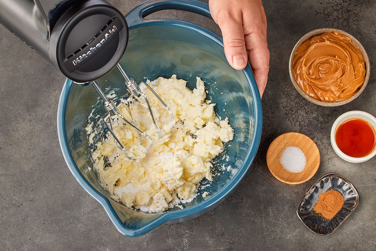 Overhead shot of a hand using an electric mixer to blend butter and sugar in a blue bowl; with small bowls of peanut butter, vanilla extract, salt, and cinnamon nearby on a gray countertop;