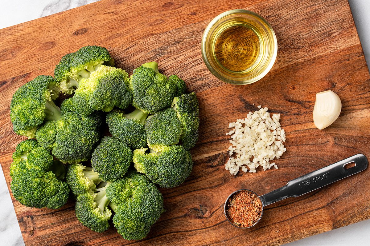 Fresh broccoli florets, a small bowl of oil, chopped garlic, a whole garlic clove, and a measuring spoon of spices are arranged on a wooden cutting board.