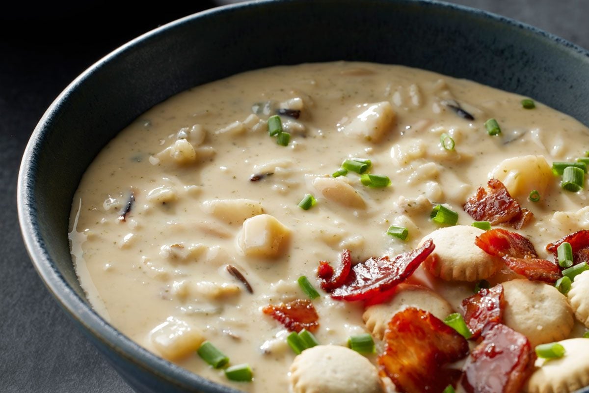 closeup shot of a creamy soup in a dark bowl, topped with chopped chives, crispy bacon pieces, oyster crackers, and small chunks of potatoes and wild rice