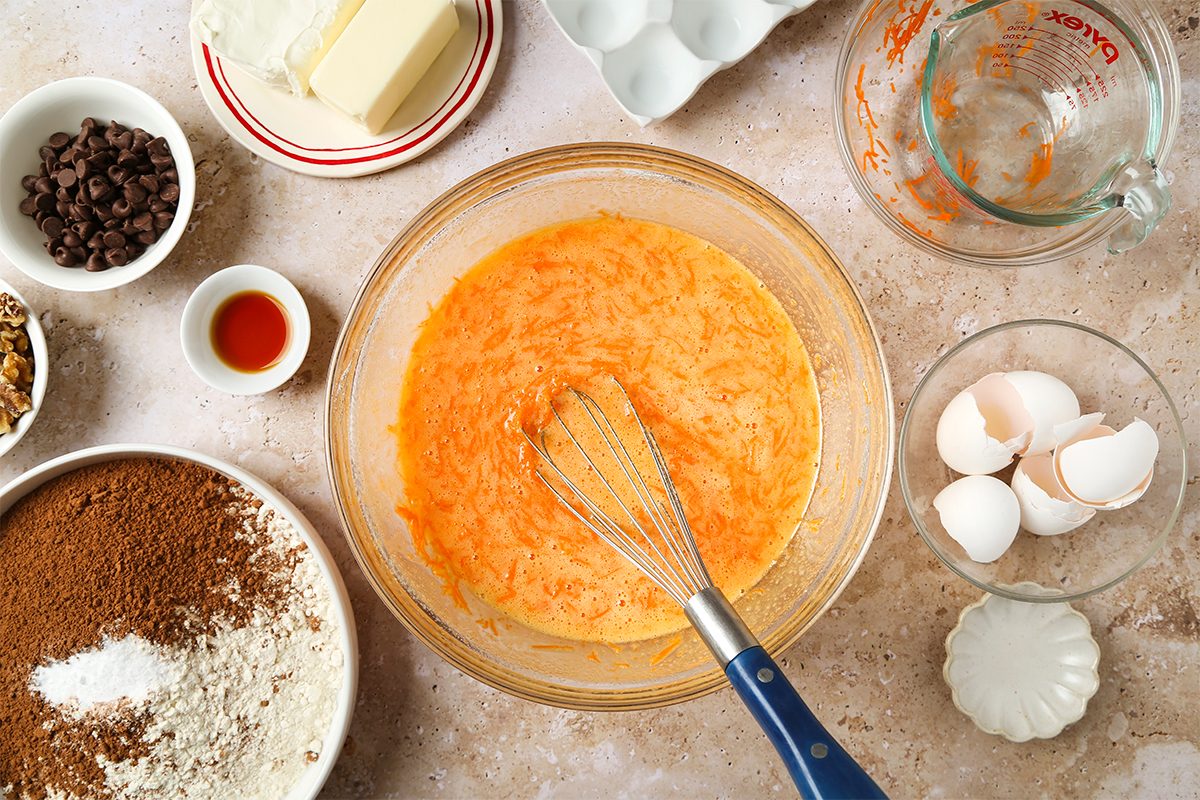 A bowl of grated carrots, eggs, and liquid being whisked, surrounded by baking ingredients: butter, chocolate chips, vanilla, flour, walnuts, brown sugar, egg shells, and a measuring cup on a countertop.