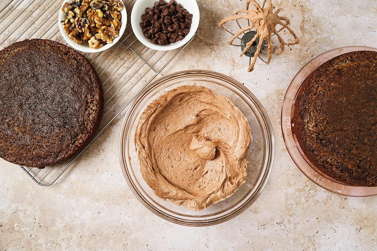Two round chocolate cake layers, a bowl of chocolate frosting, bowls of walnuts and chocolate chips, and a plastic spider are arranged on a countertop for cake decorating.