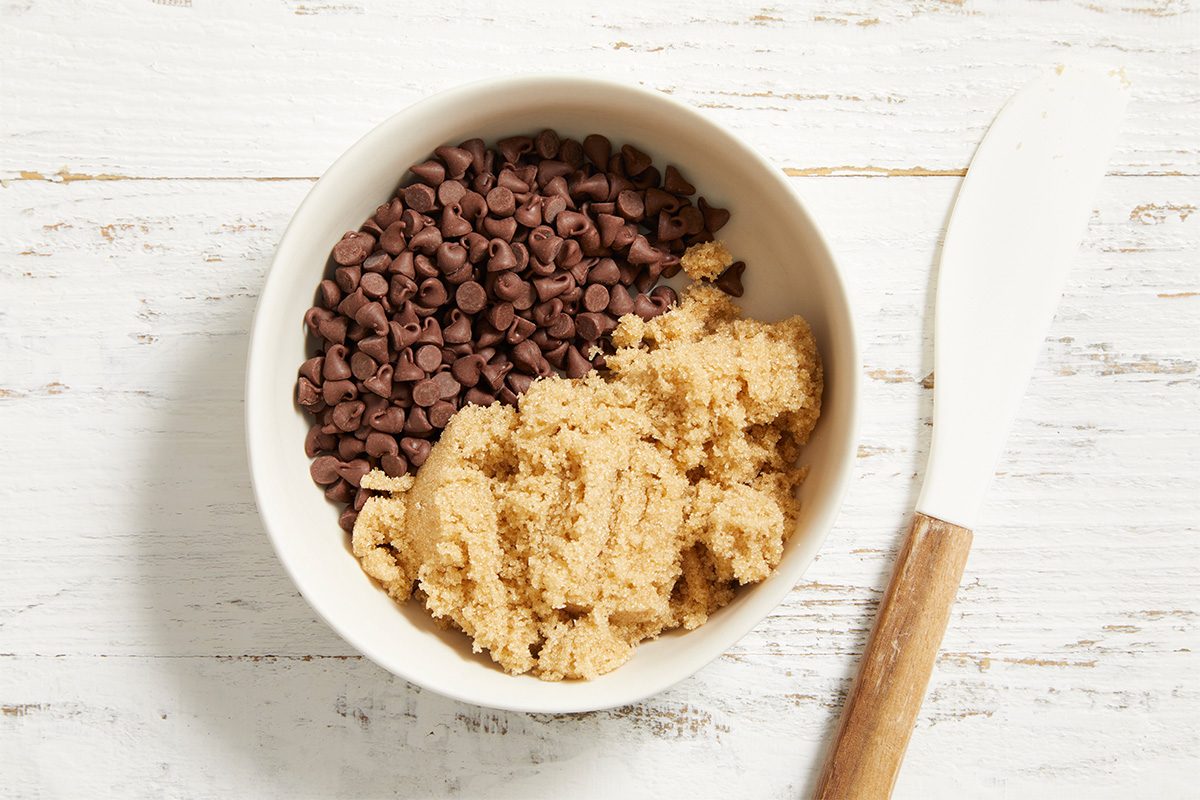 A white bowl containing chocolate chips and brown sugar sits on a white wooden surface next to a white spatula with a wooden handle.