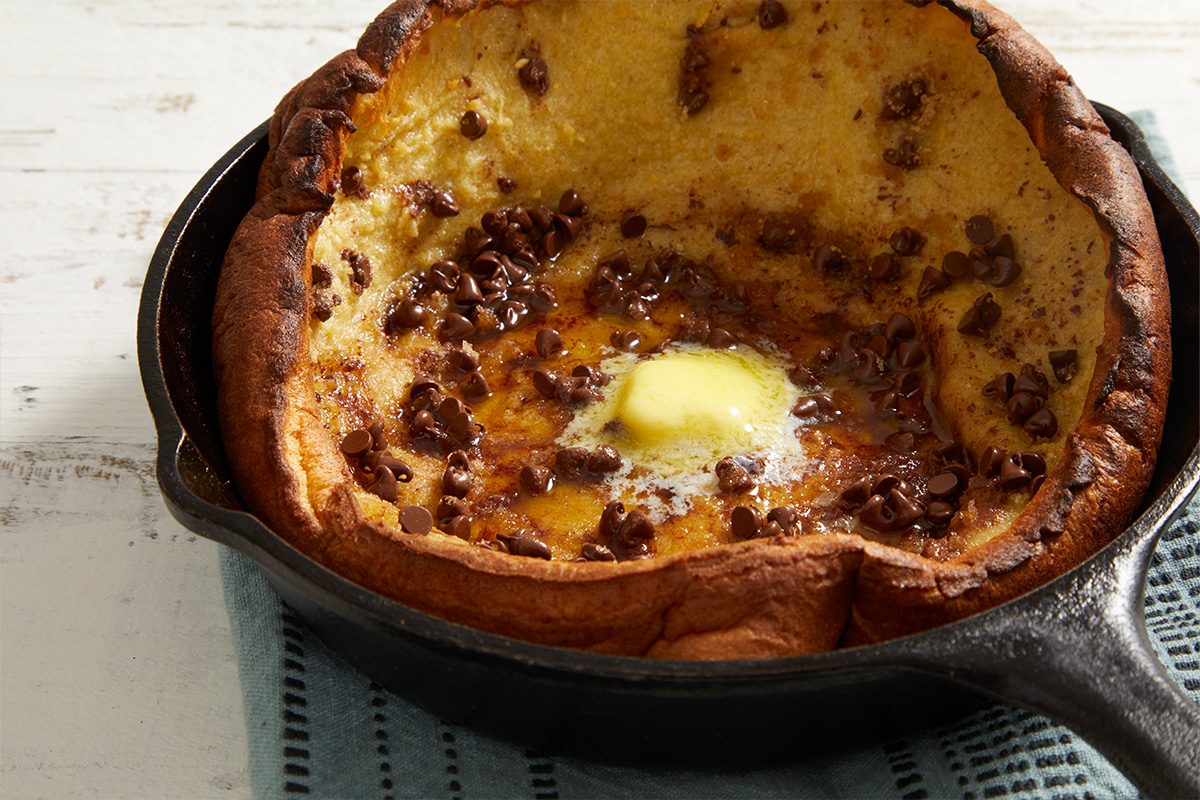 A baked Dutch baby pancake in a cast iron skillet, topped with melted butter and scattered chocolate chips, sits on a blue textured cloth on a white wooden surface.
