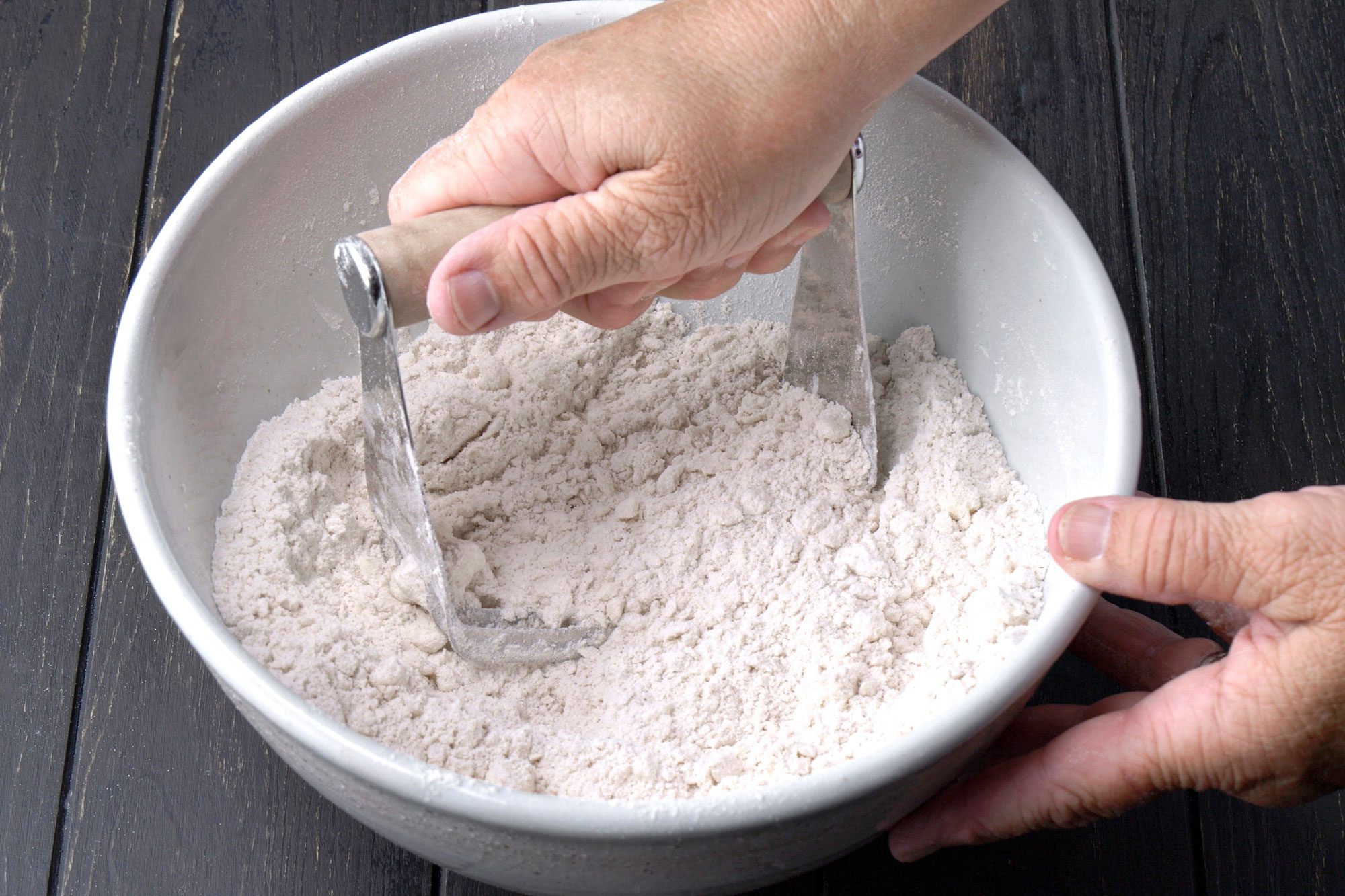 3/4th shot of a person uses a pastry blender to mix flour and fat in a white bowl on a dark wooden surface, preparing dough