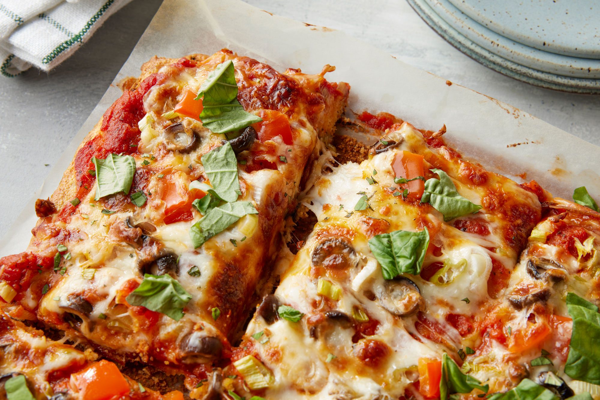 closeup shot of a rectangular veggie pizza topped with melted cheese, tomatoes, mushrooms, green onions, and fresh basil, sliced on parchment paper with plates and a napkin in the background