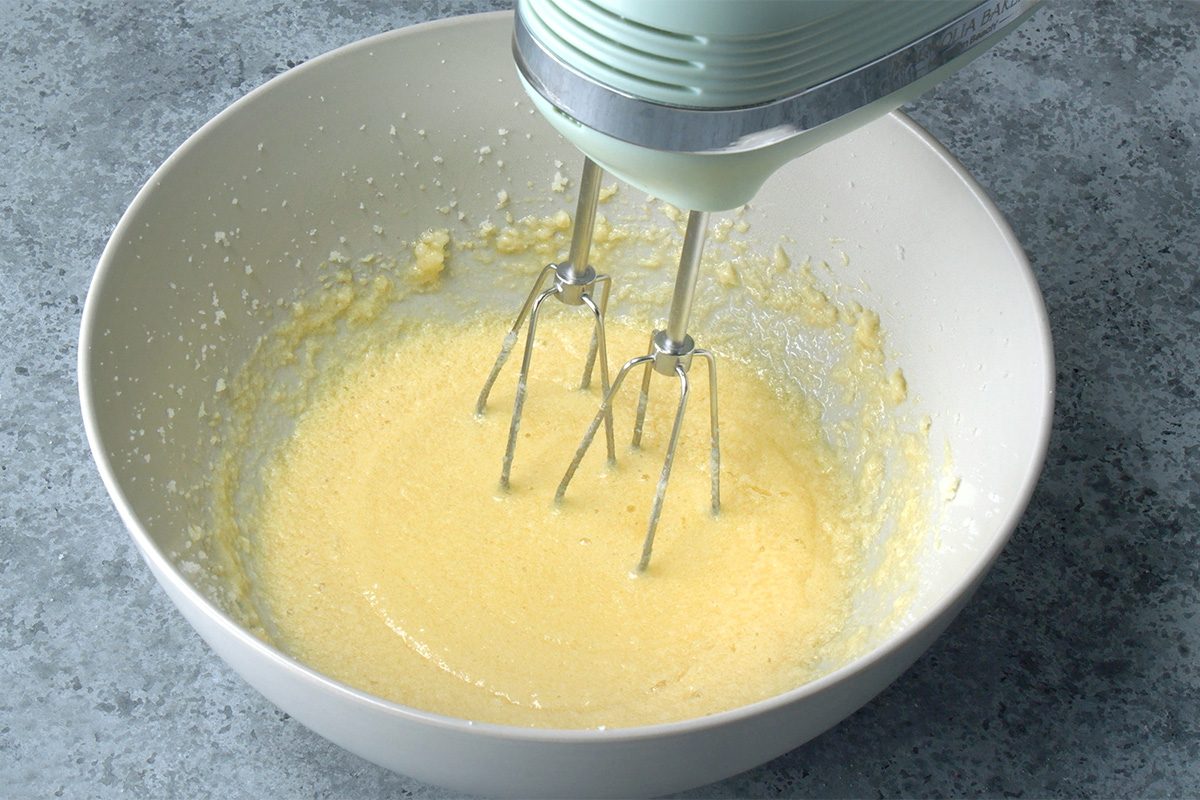 A hand mixer blends a pale yellow batter in a white mixing bowl on a gray countertop.