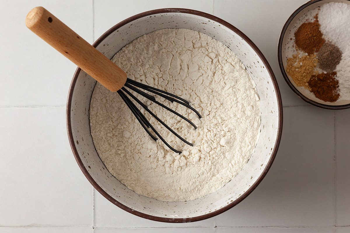 A bowl filled with flour and a whisk resting inside it, next to a smaller bowl containing assorted spices on a white tiled surface.