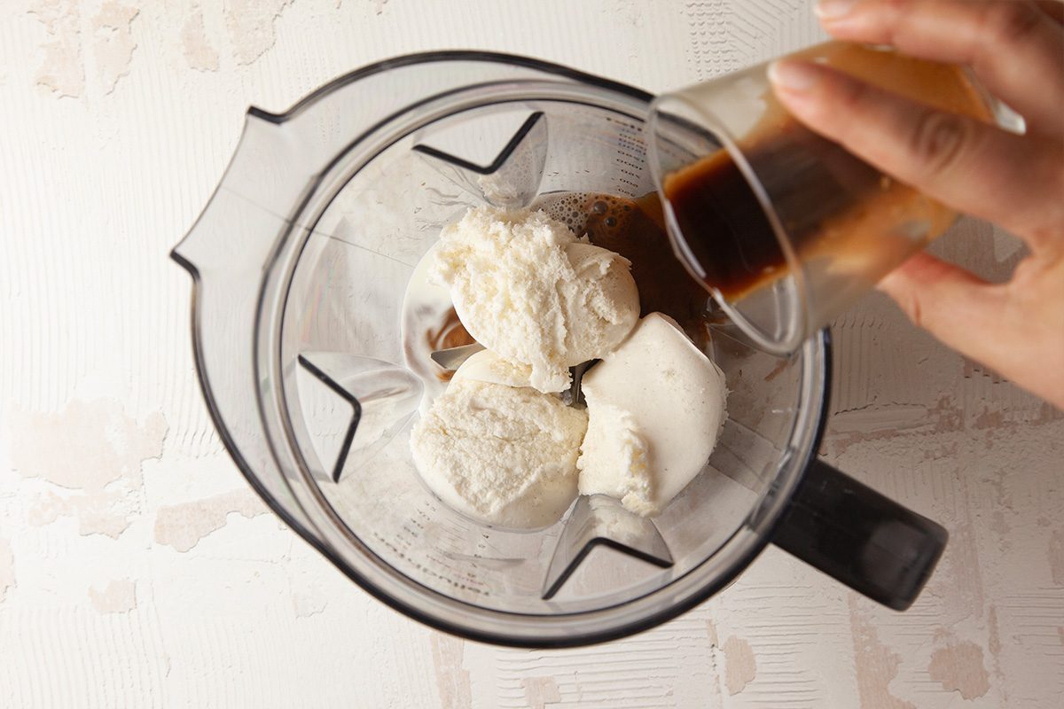 A hand pours a small cup of dark liquid, likely espresso, into a blender containing several scoops of vanilla ice cream.