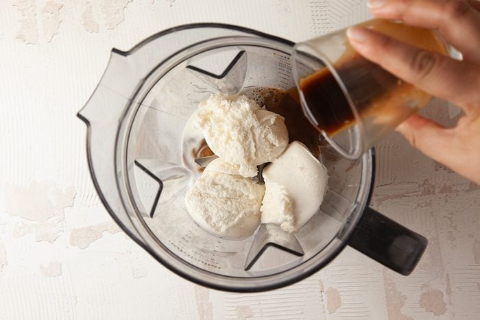 A hand pours a small cup of dark liquid, likely espresso, into a blender containing several scoops of vanilla ice cream.