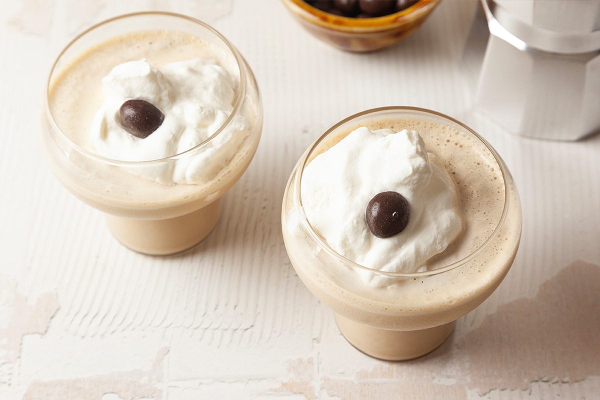 Two glass cups filled with creamy coffee topped with whipped cream and a single chocolate-covered coffee bean. A small bowl of more beans is visible in the background.