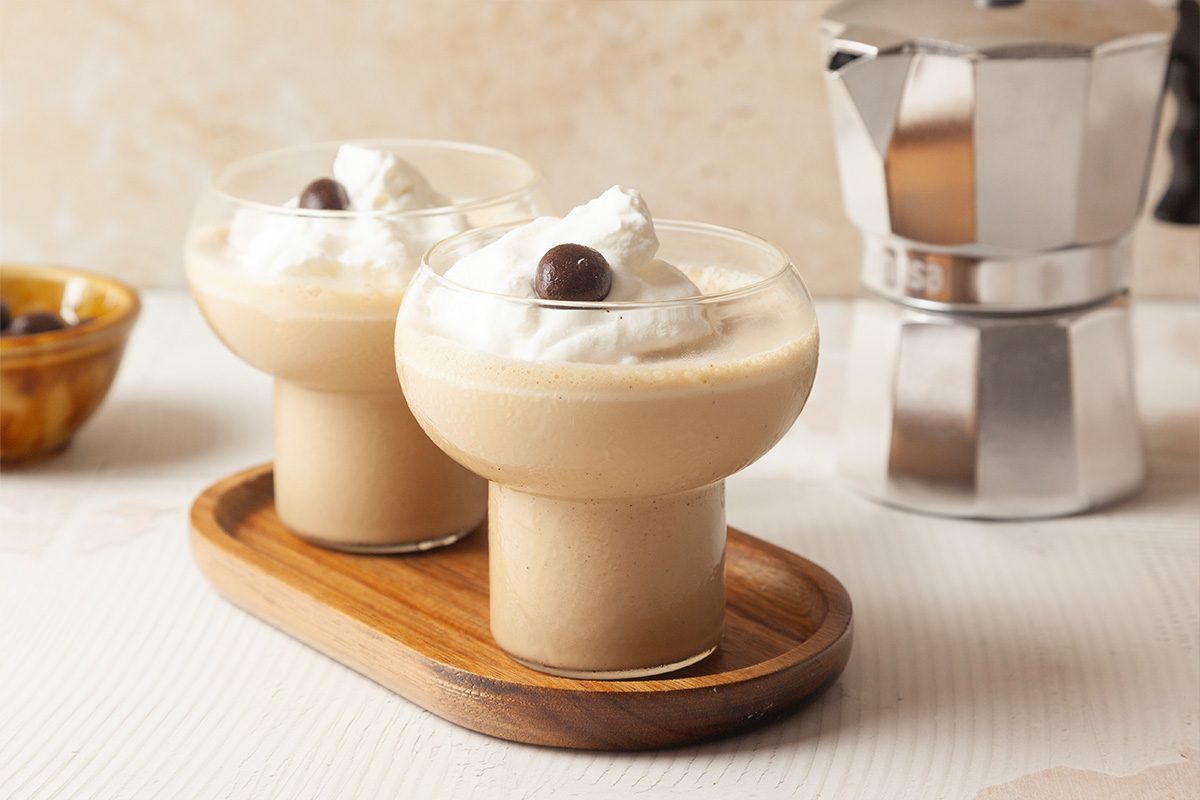Two creamy coffee drinks topped with whipped cream and a chocolate-covered coffee bean are served in round glasses on a wooden tray. A metallic coffee maker is visible in the background.