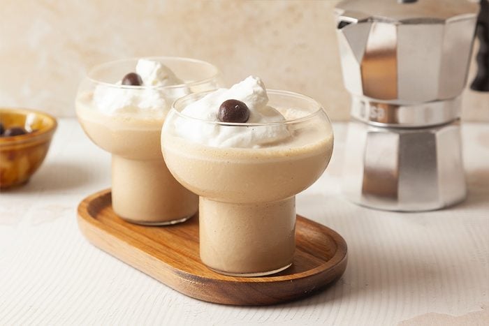 Two creamy coffee drinks topped with whipped cream and a chocolate-covered coffee bean are served in round glasses on a wooden tray. A metallic coffee maker is visible in the background.