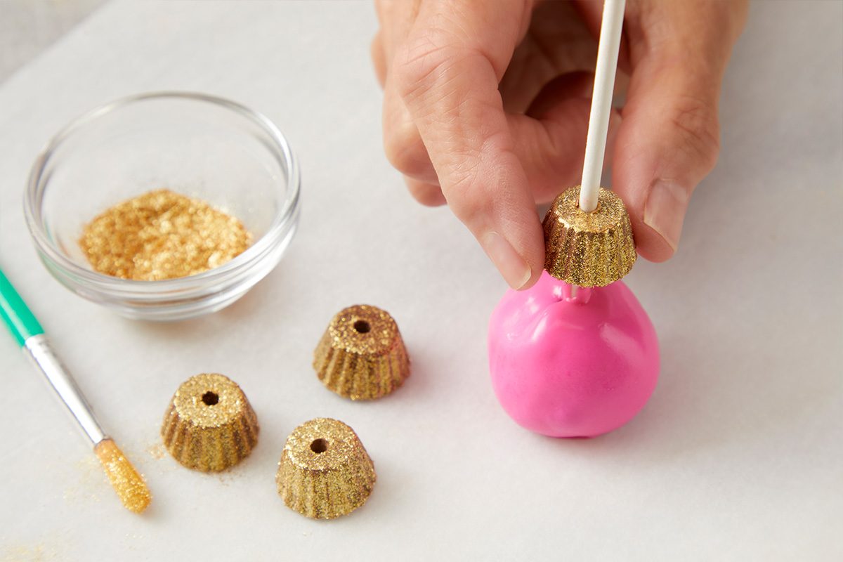 A hand attaches a gold-wrapped chocolate to a pink candy ball on a stick. Nearby are more gold chocolates, a small bowl of gold sprinkles, and a green-handled brush.