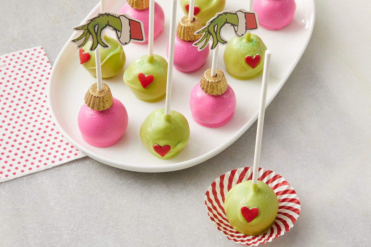 A plate of pink and green cake pops with white sticks, decorated with red hearts. Some green cake pops have Grinch hand cutouts on the sticks. One cake pop sits in a red striped paper liner beside the plate.
