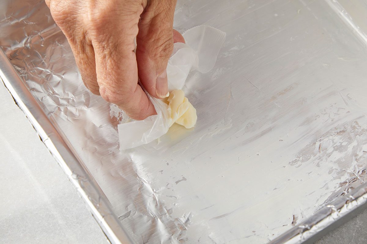 overhead shot of a hand uses a piece of wax paper to spread butter on the surface of a foil lined baking pan