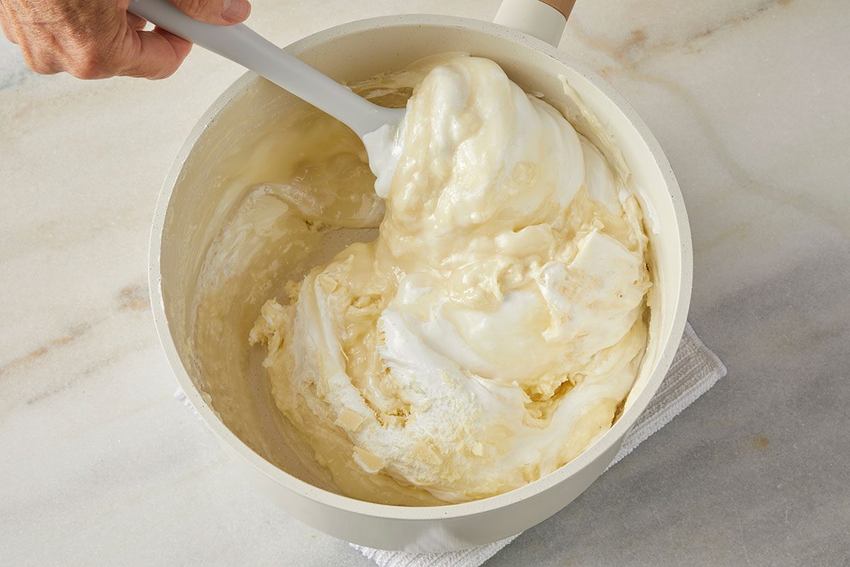overhead shot of a hand stirs a thick, creamy mixture in a white saucepan with a white spatula; The pot sits on a white cloth on a marble countertop