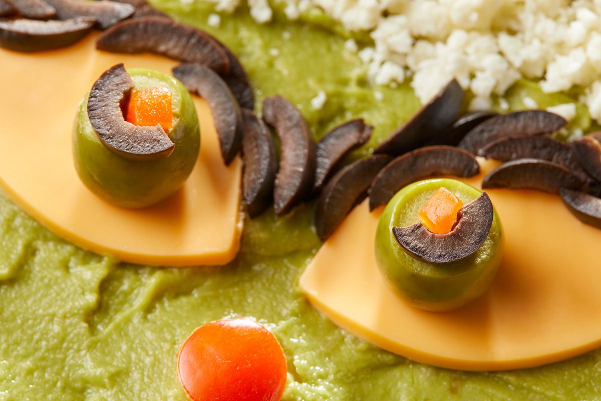 Close up shot of a festive guacamole dish decorated with cheese slices; green olives; black olive slices; diced red pepper; and crumbled cheese; arranged to resemble faces with eyes