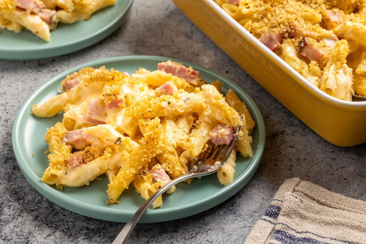 Overhead shot; plate of baked macaroni and cheese with ham; crispy breadcrumb topping; casserole dish; fork; striped cloth napkin