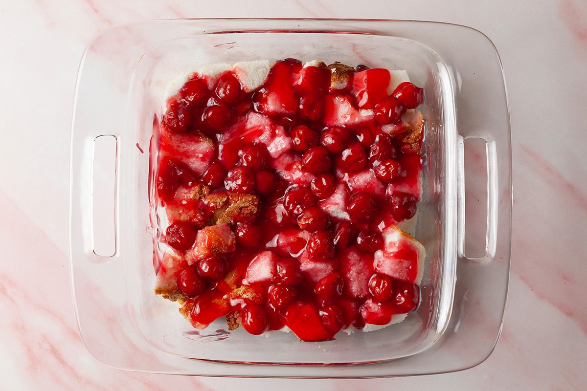 A glass baking dish filled with a dessert topped with bright red cherry pie filling and cream layers, set on a light pink marble surface.