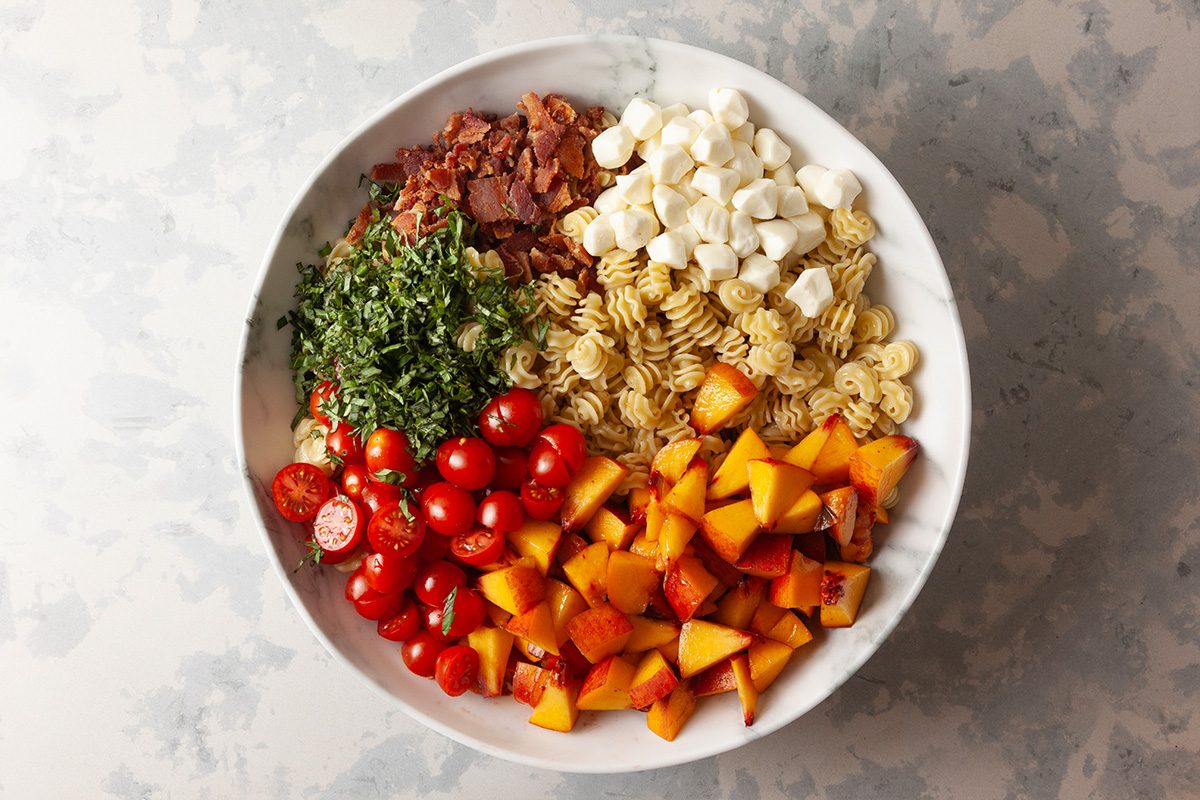 A white bowl filled with sections of pasta, cherry tomatoes, roasted butternut squash, chopped basil, mini mozzarella balls, and crumbled bacon, arranged neatly on a light textured surface.