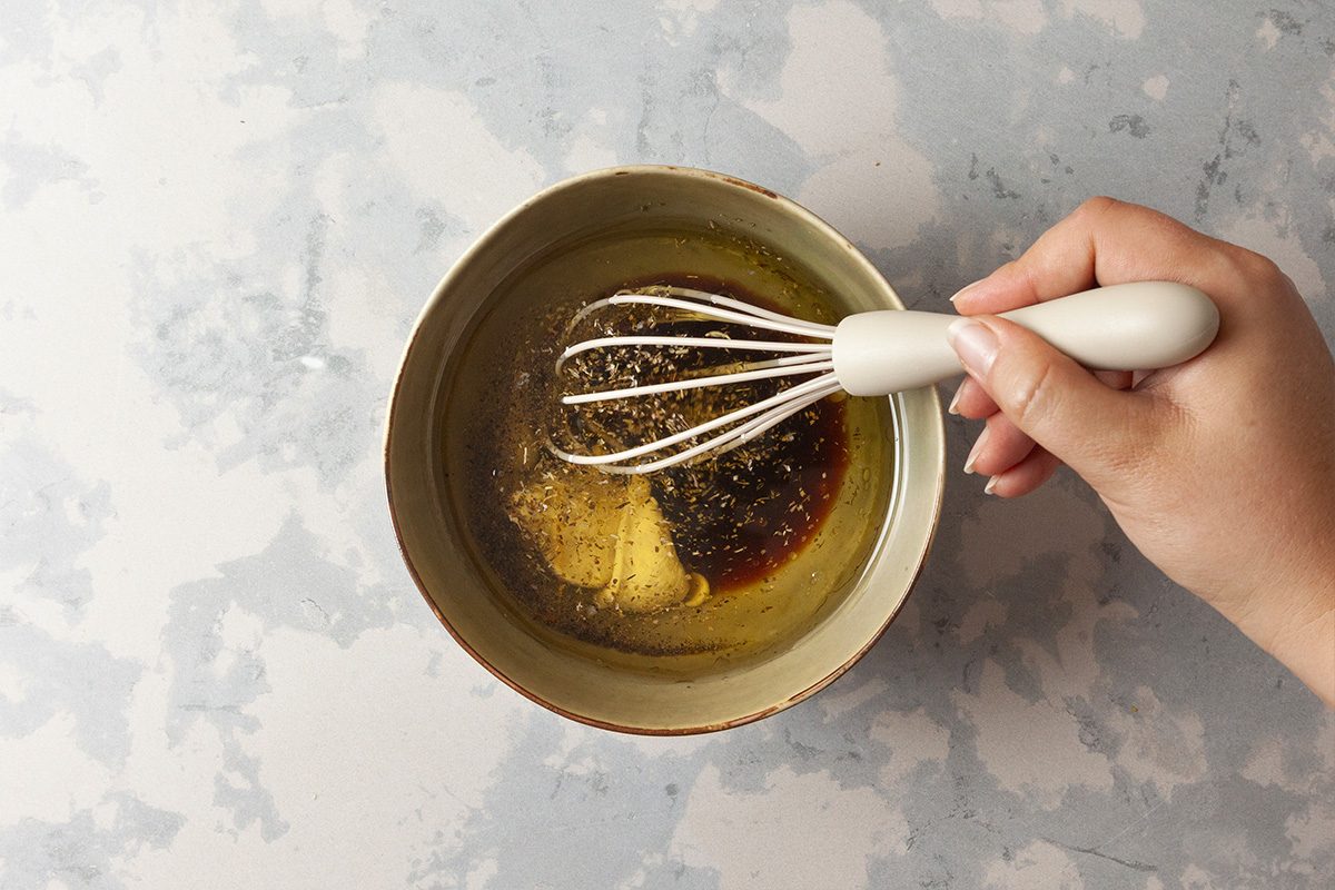 A hand holds a whisk mixing oil, mustard, and spices in a bowl on a light, marbled surface.