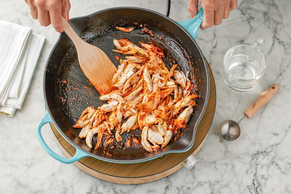 This is a close-up kitchen shot; a person stirs shredded, seasoned chicken in a blue skillet on a marble countertop; nearby are a folded towel; glass water pitcher; and metal measuring cup