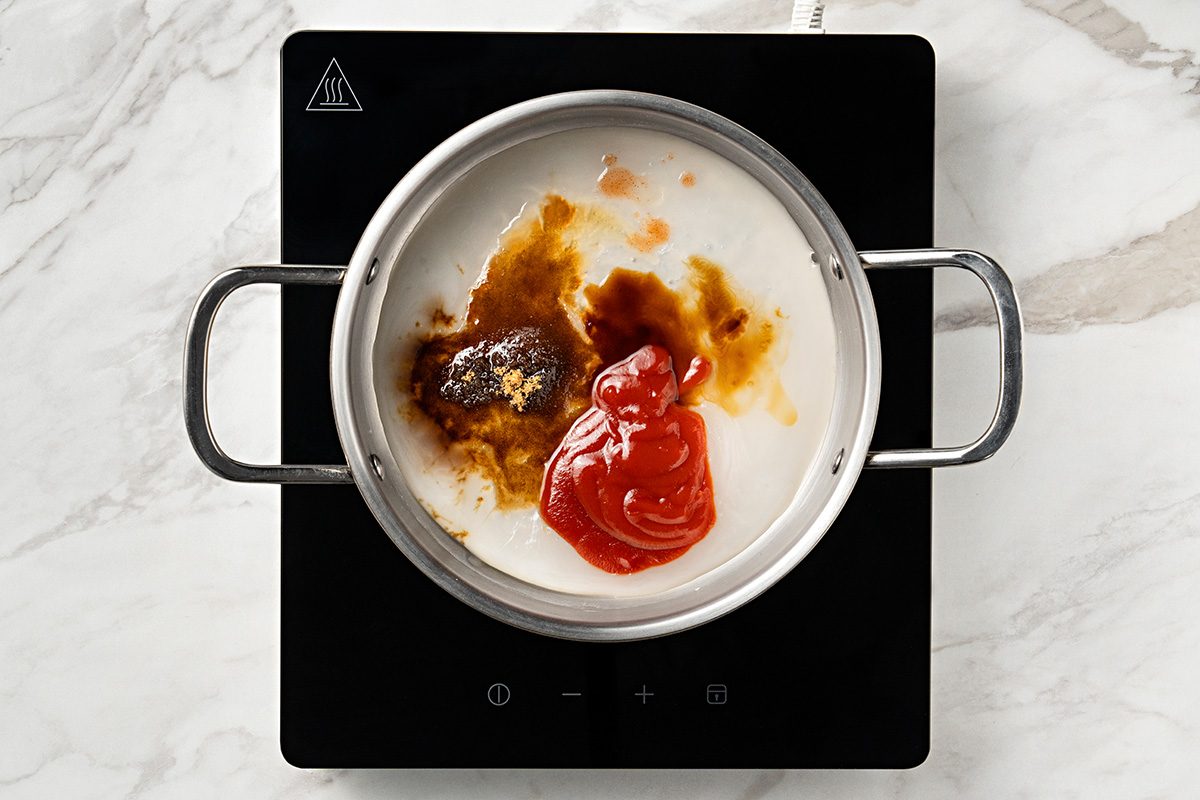 A saucepan on an electric stovetop contains milk, ketchup, soy sauce, and seasoning, viewed from above on a marble countertop.