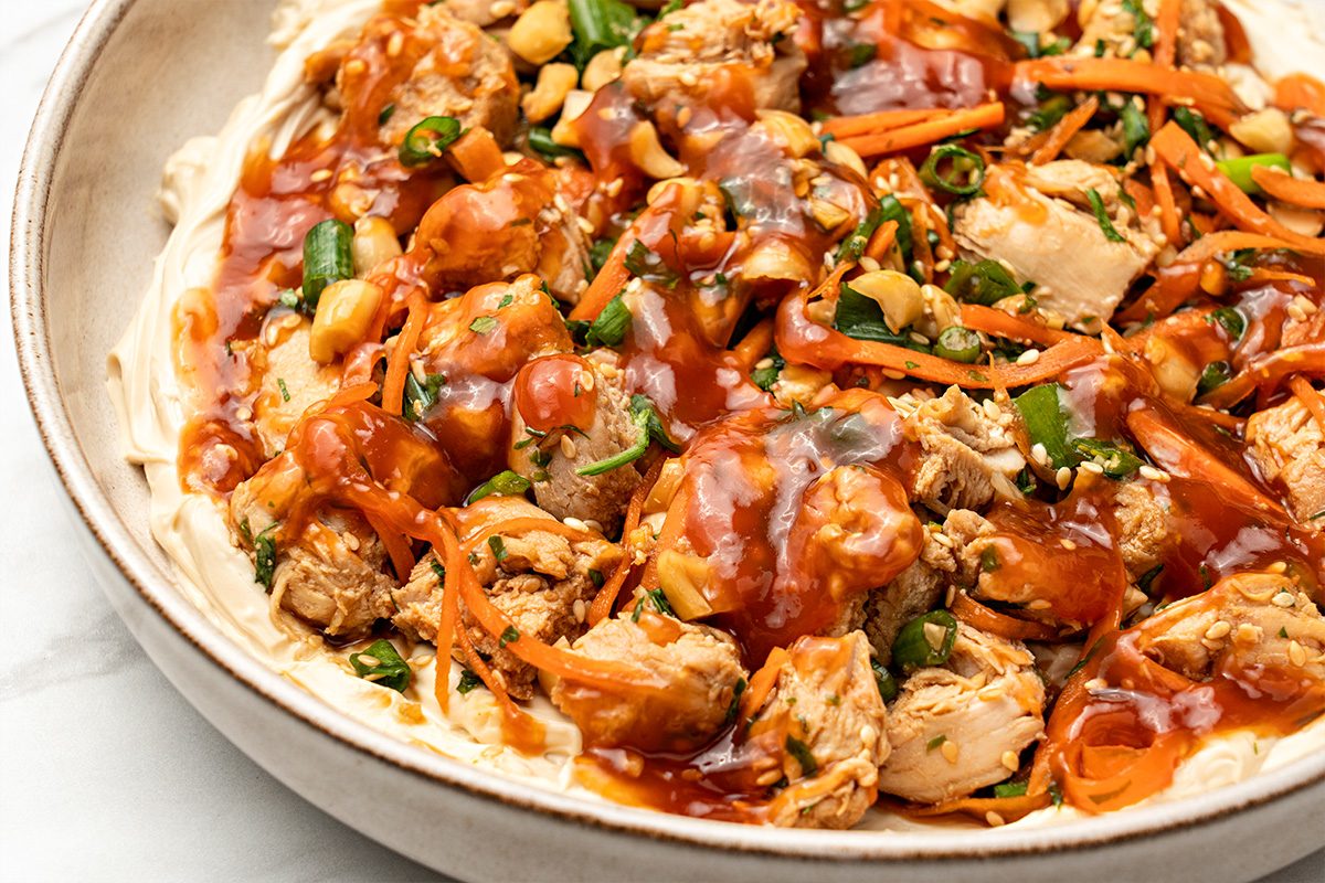 Close-up of a bowl filled with shredded chicken, sliced green onions, carrots, peanuts, and a red sauce on a creamy base. The dish appears to be garnished with sesame seeds and herbs.