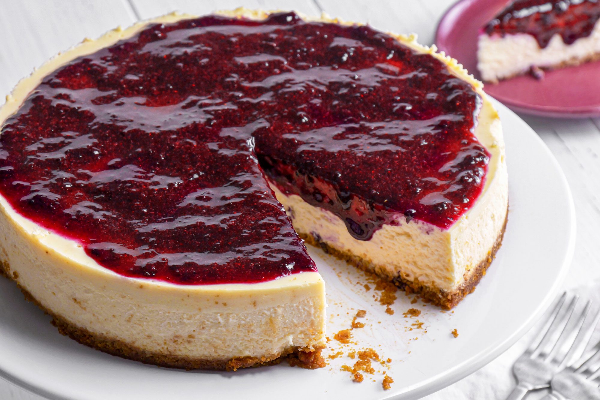 3/4th shot of a Lemon Blueberry cheesecake topped with glossy berry sauce sits on a white plate, with a single slice removed and placed on a red plate nearby, Three forks are visible on a white napkin in the corner