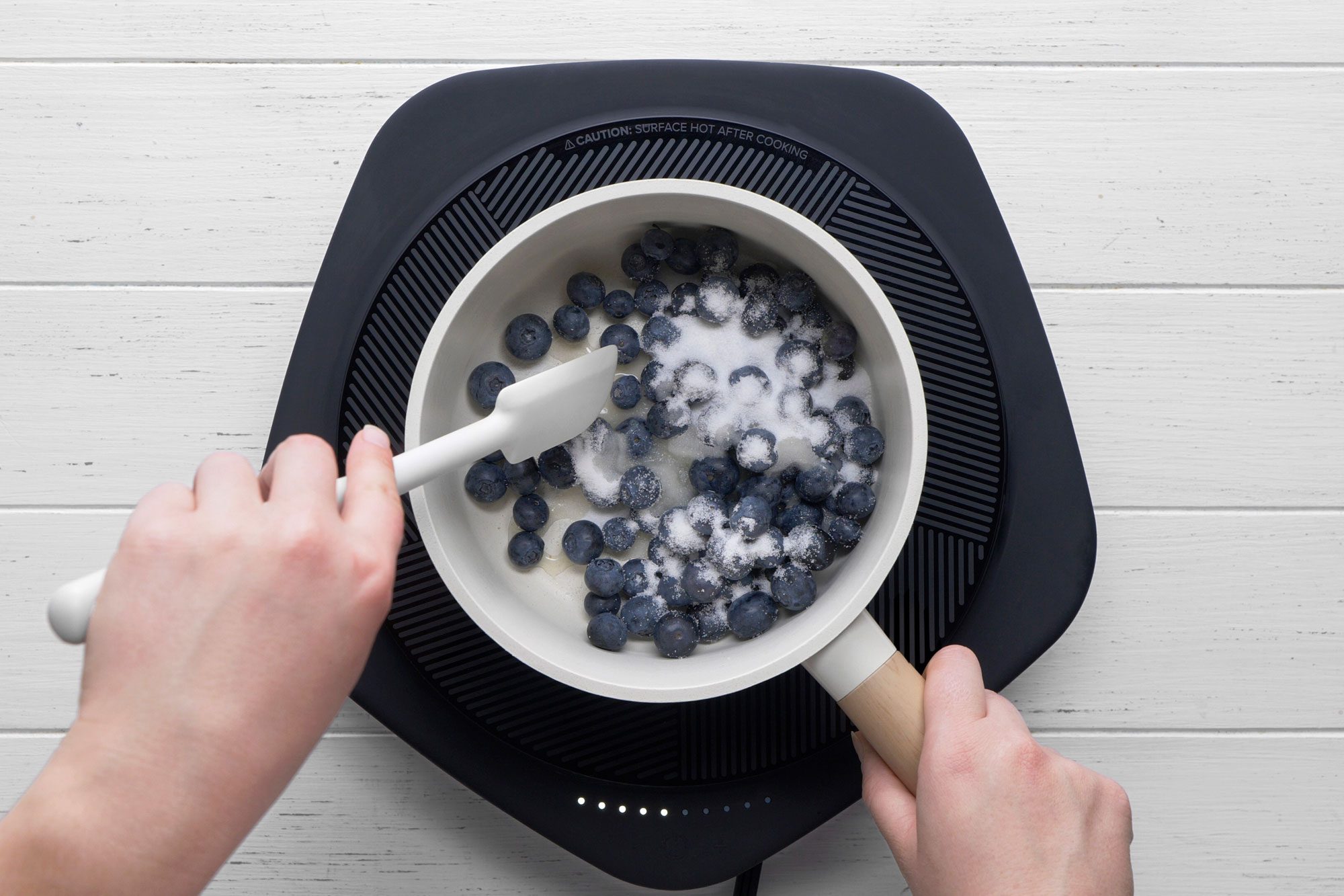 overhead shot of a person stirs blueberries and sugar in a white saucepan on an electric cooktop, preparing a mixture on a white wooden surface