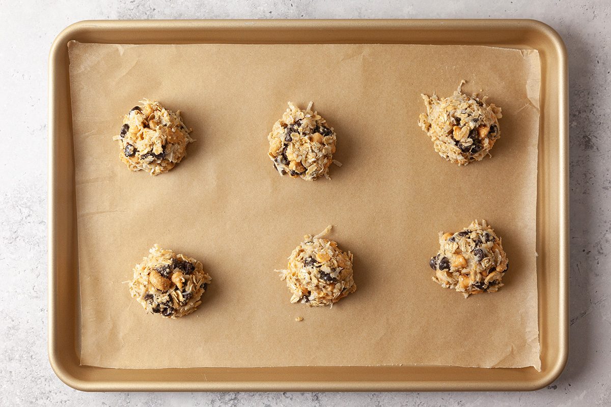 A baking sheet lined with parchment paper holds six evenly spaced scoops of unbaked cookie dough with visible chocolate chips.