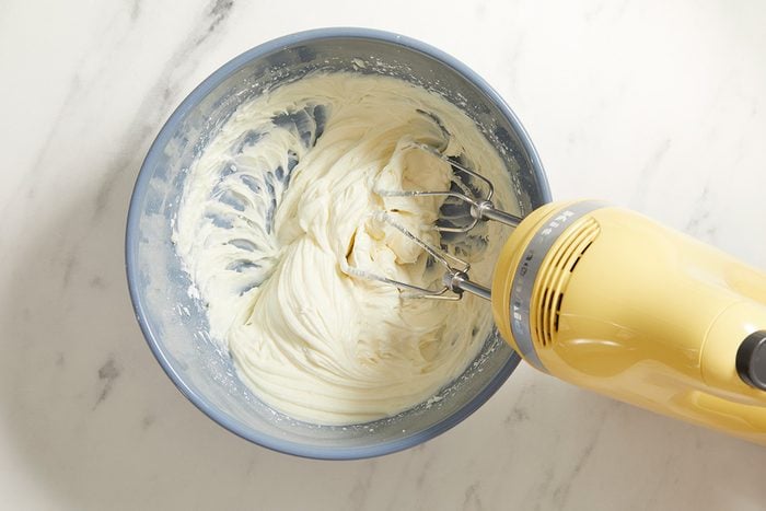 A yellow electric hand mixer is blending a creamy white mixture in a blue bowl on a white marble surface.